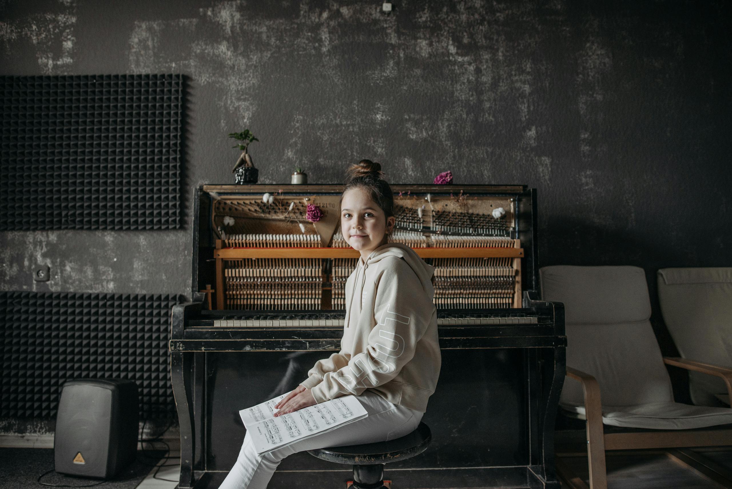 A young girl sits smiling at an upright piano, holding a music sheet. Perfect for music education themes.