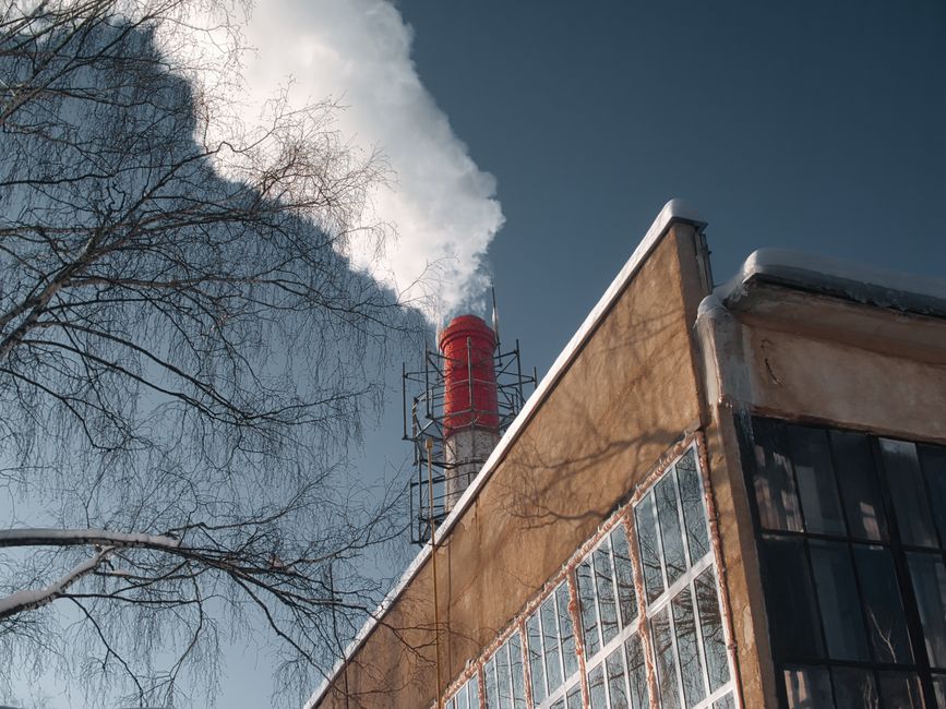 A factory chimney emits white smoke, casting a shadow on a clear day beside a bare winter tree.