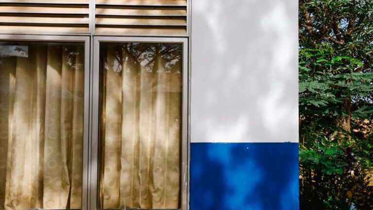 Close-up of a building facade with windows and beige curtains, reflecting trees outside.